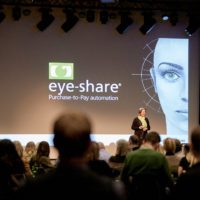 Woman (Dina Drehn) talking on stage in front of audience. A big screen with the logo of eye-share is in the background
