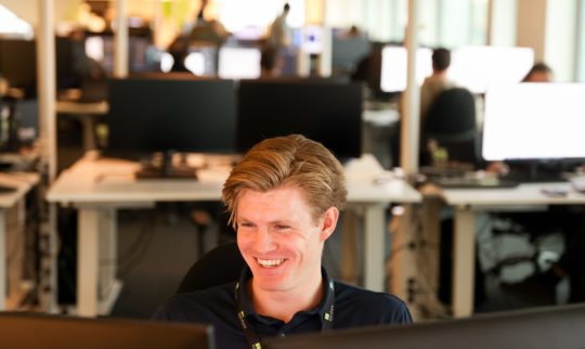 Male eye-share employee in office environment, sitting in front of computer smiling.