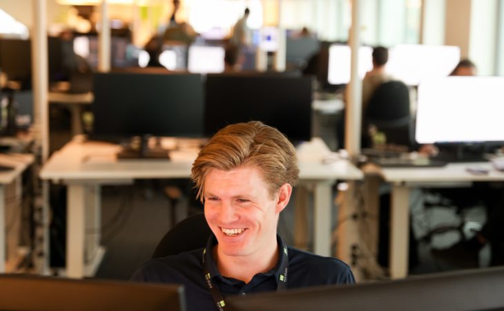 Male eye-share employee in office environment, sitting in front of computer smiling.