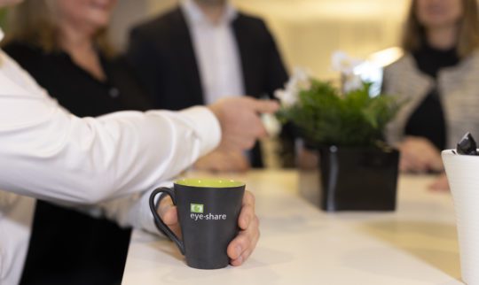 Black branded eye-share coffee mug with green eye-share logo placed on table in office environment.