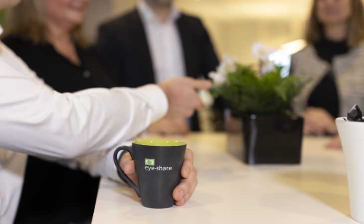 Black branded eye-share coffee mug with green eye-share logo placed on table in office environment. Group of people blurred out in the background.