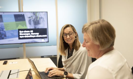 Eye-share employees Torhill and Siw talking in a meeting room sitting in front of their laptops.